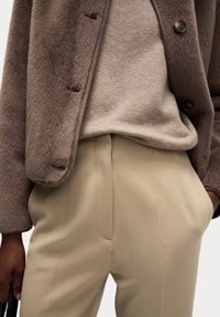 Brun pälsjacka i syntetmaterial över en beige tröja och krämfärgade avslappnade byxor. Anmärkningsvärda sömnadsdetaljer och mjuka texturer genom hela outfiten.