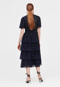 Vestido de color azul marino con volantes y mangas cortas, adornado con lentejuelas. Presenta un corpiño ajustado y una falda fluida, acompañado de sandalias de tacón transparente.