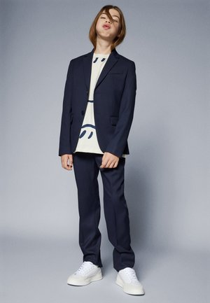 Completo blu navy con blazer a due bottoni, pantaloni abbinati e una t-shirt crema con un design di un sorriso nero. Sneakers bianche completano il look.