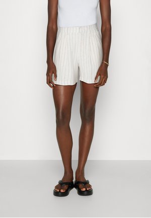 Personne vêtue d'un short blanc taille haute avec de fines rayures verticales, d'un haut blanc sans manches, de sandales compensées noires, debout devant un fond blanc.