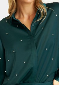 Smaragdgroene shirt met een knoopsluiting, met kleine kristalversieringen verspreid over de stof voor extra detail.