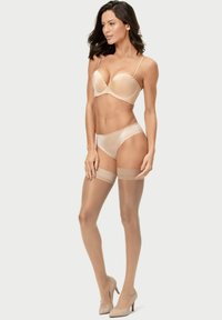 Nackter BH mit gewellten Kanten, passenden Panties und transparenten, hüfthohen Strümpfen. Das Model trägt beigefarbene High Heels, die eine glatte Silhouette betonen.