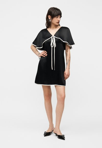 Γυναίκα που στέκεται, φορώντας ένα κοντό μαύρο σατέν φόρεμα με λευκή περιφέρεια, γιακά σε σχήμα κάπας και μαύρα μυτερά mule με αλυσίδες.