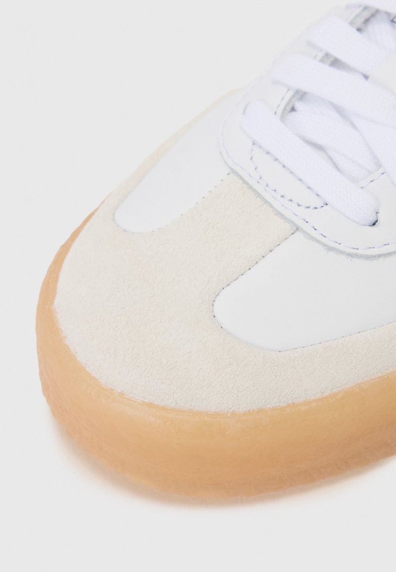 Sneaker bianco con tomaia in morbida pelle scamosciata e suola in gomma. Presenta un design con lacci e una superficie liscia. Contrasto notevole tra i materiali.