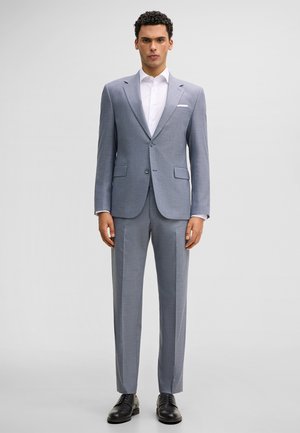 Mann steht, trägt einen hellgrauen Zweiknopfanzug mit weißem Hemd, Einstecktuch und schwarzen Anzugschuhen, vor schlichtem hellem Hintergrund.