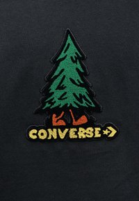 Το μαύρο  ύφασμα διαθέτει σχέδιο πράσινου δέντρου με πορτοκαλί λεπτομέρειες στη βάση, συνοδευόμενο από το κείμενο "CONVERSE" σε κίτρινο χρώμα με ένα βέλος προς τα δεξιά.