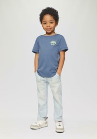 Blauwe katoenen T-shirt met korte mouwen, voorzien van een groene dinosaurustekening. Lichtblauwe jeans met voorzakken en witte sneakers met grijze accenten.