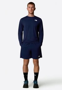 Μπλούζα με μακριά μανίκια σε ναυτικό χρώμα και σορτς από ελαφρύ ύφασμα. Διαθέτει το λογότυπο της The North Face σε λευκό χρώμα. Το μοντέλο φοράει σκούρες κάλτσες και αθλητικά παπούτσια.