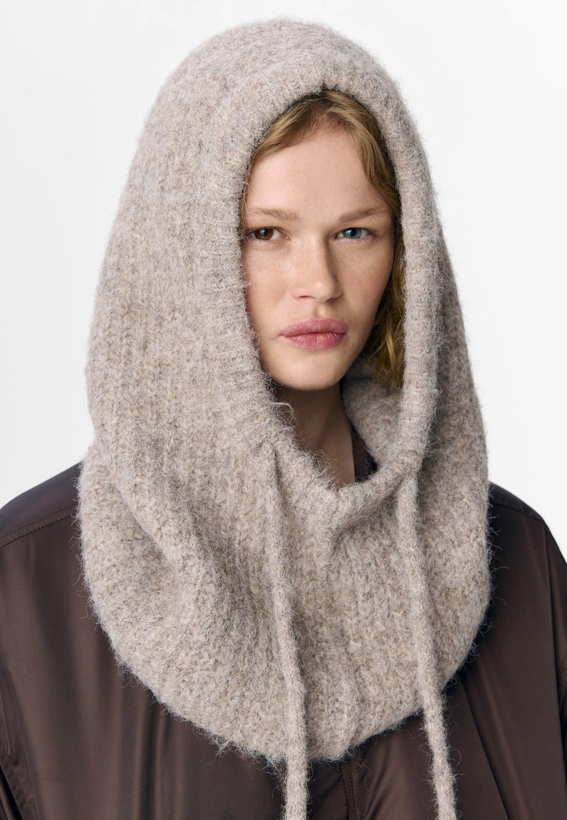 Beige knit hood made from soft, textured wool with a slouchy design, featuring two ties and a smooth inner lining.