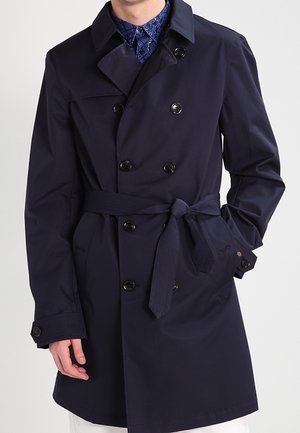 Marineblauer doppelt geknöpfter Trenchcoat mit Gürtel in der Taille gebunden, getragen über einem gemusterten blauen Hemd, mit einer Hand in der Tasche.
