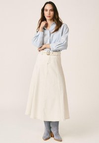 Femme portant un chemisier bleu clair à motifs, rentré dans une jupe crayon crème taille haute avec ceinture et bottines chevilles en daim gris.