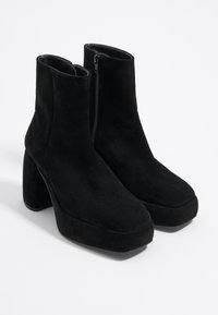 Botines de ante negro con tacón grueso y suela de plataforma. Cuentan con un cierre lateral y una textura suave y mate. Diseño minimalista.