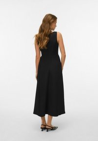 Robe sans manches noire avec un corsage ajusté, jupe évasée fluide et détails de coutures subtils ; associée à des escarpins noirs à motifs.