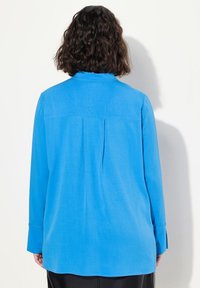 Camicia blu a maniche lunghe realizzata in tessuto morbido, con piega sul retro, polsini con bottoni e colletto classico. Orlo dritto con bordi arrotondati.
