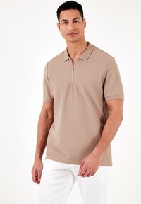 Bežs īsām piedurknēm polo krekls ar ceturtdaļas rāvējslēdzēju, strukturētu audumu un klasisku apkakli. Apvienots ar baltām biksēm.