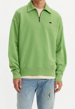 Sweat-shirt vert à fermeture éclair avec col, doté de poignets côtelés et d'un petit logo sur la poitrine. Porté par-dessus un tee-shirt de couleur claire et un jean.