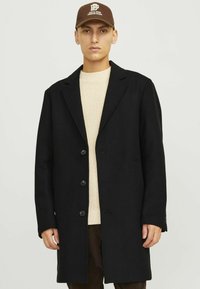 Abrigo de lana negro con solapas en pico, tres botones y bolsillos laterales, combinado con un suéter texturizado crema y una gorra marrón.