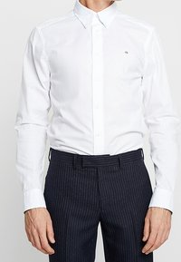 Witte overhemd met knopen en een spitse kraag met een klein logo. Draagt donkere broek met krijtstreep, die een getailleerde pasvorm laat zien.