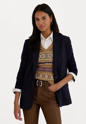 Blazer rayé bleu marine sur un gilet en tricot multicolore, chemise blanche en dessous et pantalon marron avec une ceinture à boucle en argent.