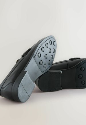 Schwarze Schuhe mit einem glatten Lederobermaterial und einer strukturierten Gummisohle, die runde Grifffelder aufweist. Die Sohle hat eine auffällig robuste Konstruktion.