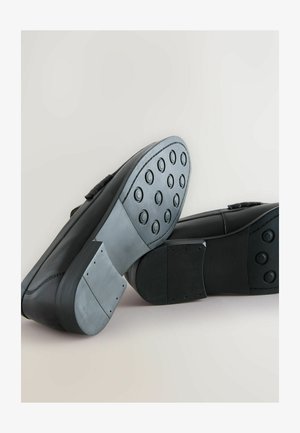 Schwarze Schuhe mit einem glatten Lederobermaterial und einer strukturierten Gummisohle, die runde Grifffelder aufweist. Die Sohle hat eine auffällig robuste Konstruktion.