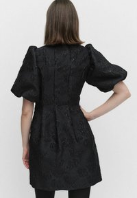 Svart klänning med puffärmar, texturerat tyg med blommönster, figurnära midja och en utsvängd kjol. Visad bakifrån.