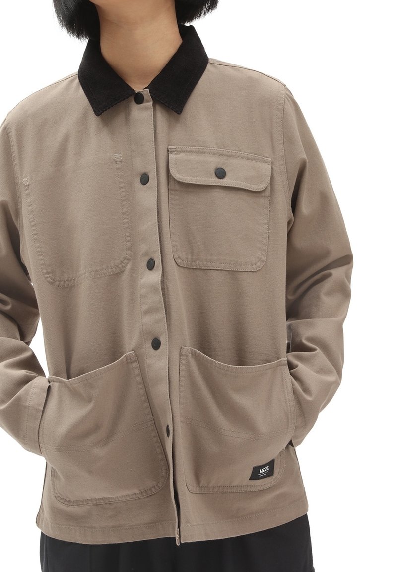 Personne portant une veste beige à boutons avec un col noir et plusieurs poches avant, les mains dans deux poches inférieures.