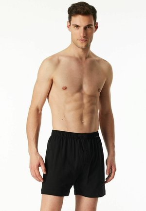 Young man with short dark hair wearing black boxer shorts, standing against a plain white background, facing forward with a neutral expression.