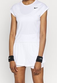 Top deportivo blanco de manga corta, combinado con una falda blanca. Logo negro de Nike en el top y muñequeras negras. Tejido suave y texturizado.