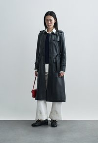 Trenci din piele neagră cu nasturi în față, talie cu curea, buzunare laterale, purtat peste un pulover bleumarin, asortat cu pantaloni largi crem și pantofi negri.