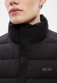 Μαύρο μπουφάν με επένδυση και ψηλό γιακά. Διαθέτει ομαλή υφή και αντιθετικό οριζόντιο μοτίβο με ραφές. Το λογότυπο "BOSS" είναι τυπωμένο στον ώμο.