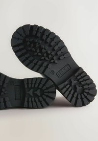 Semelles de chaussures en caoutchouc noir avec des motifs de semelle profonde, dotées de crampons hexagonaux et triangulaires pour une meilleure adhérence. Nom de la marque "NEXT" gravé sur le côté.