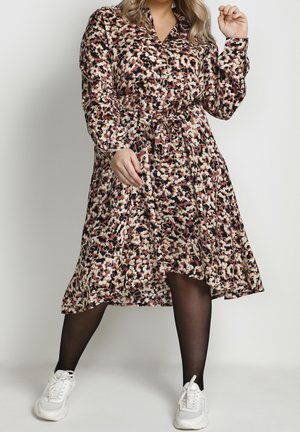 Femme portant une robe midi à manches longues aux motifs marron, noir et blanc avec ceinture à la taille, collants noirs et baskets blanches, debout jambes écartées.