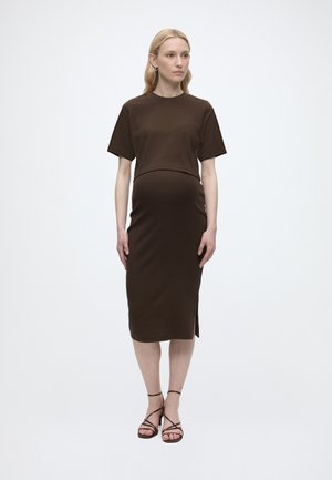 Rase naine, kes kannab tumedat pruuni soonikkoes lühikeste varrukatega kleiti ja rihmikuid kontsakingi, seisab tavalise heleda tausta ees.