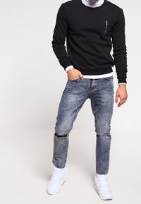 Sudadera negra de cuello redondo con puños y dobladillo acanalados, con un bolsillo en el pecho con cremallera. Vaqueros azul claro desgastados con detalle de rodilla rota. Zapatillas blancas.