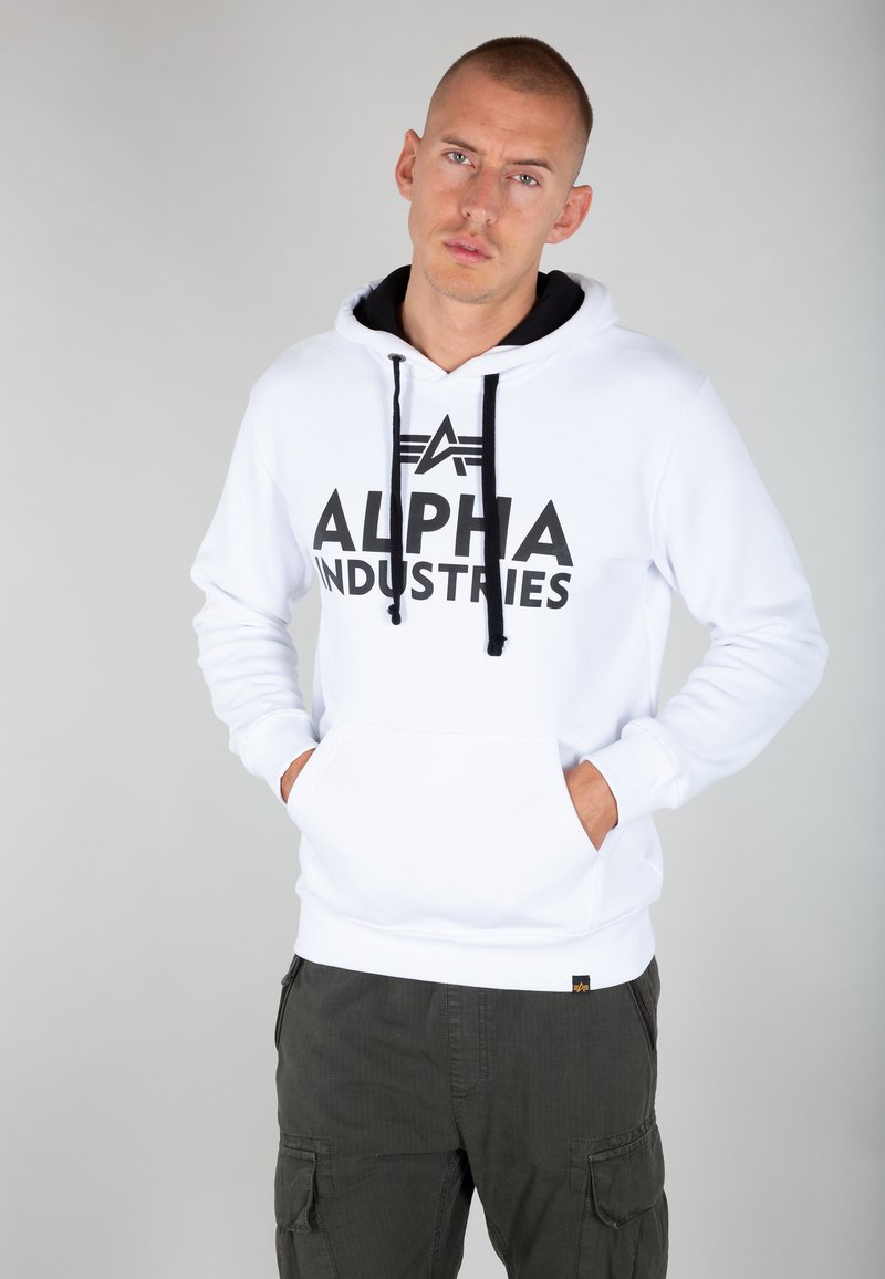 Sudadera blanca con cordones negros y "ALPHA INDUSTRIES" impreso en grandes letras negras en la parte delantera; cuenta con un bolso tipo canguro.