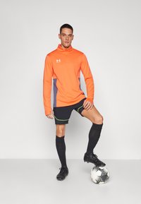 Tenue de sport comprenant un haut zippé à manches longues orange avec des accents gris, un short noir, des chaussettes noires et des chaussures de football, positionnée à côté d'un ballon de soccer.
