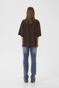 Person with straight brown hair wearing a loose dark brown top and faded blue jeans, standing facing away on a plain white background.