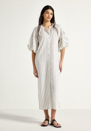 Kvinde stående iført en hvid midikjole med tynde lodrette striber, puffede trekvartærmer og sorte flade sandaler.