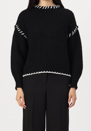 Schwarzer Strickpullover mit Rippstruktur, weißen Akzenten am Kragen und Saum sowie weiten Ärmeln. Kombiniert mit schwarzen Hosen.