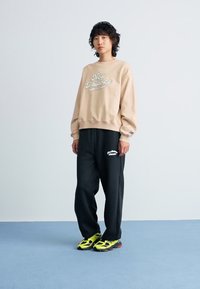 Sweatshirt beige avec le logo "New Balance", associé à un pantalon de survêtement noir avec des imprimés blancs. Des sneakers jaune vif et noir complètent le look.