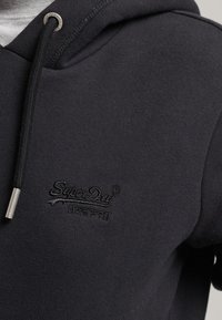Felpa nera con cappuccio e cordino, dotata di dettagli metallici argentati. Presenta un logo ricamato "SuperDry" sul petto. Tessuto morbido e testurizzato.