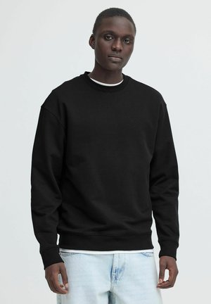 Giovane uomo che indossa una felpa nera a maniche lunghe sopra una camicia bianca e jeans azzurri, in piedi davanti a uno sfondo chiaro e uniforme.