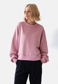 Różowy sweatshirt z okrągłym dekoltem, luźny krój i obniżone ramiona. Miękka faktura, szerokie mankiety i prążkowany dół w zestawieniu z czarnymi spodniami.