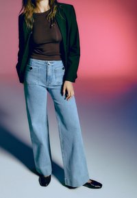 Femme portant un blazer noir, un haut marron, un jean large bleu clair avec des poches avant, et des chaussures noires, debout devant un fond dégradé rose.