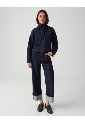Vrouw die een donkerblauwe spijkerjasje en broek met brede boorden draagt, donker coltrui en zwarte loafers, staand tegen een effen achtergrond.