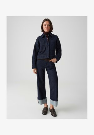 Frau trägt dunkelblaue Jeansjacke und Hose mit weiten Bündchen, dunklen Rollkragen und schwarzen Loafers, steht vor einfarbigem Hintergrund.