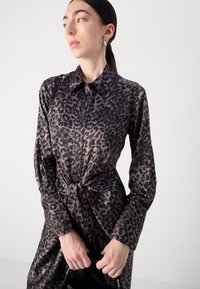 Abito camicia in satin con stampa leopardata in nero e marrone. Presenta una chiusura con bottoni, colletto e dettagli con cintura in vita, con maniche lunghe.