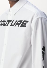 Weißes Hemd mit Knopfleiste, auf der Brust groß das schwarze "COUTURE"-Logo und schwarze Akzente an den Ärmeln. Glatter Stoff mit langen Ärmeln.