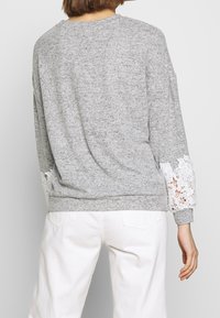 Grå sweatshirt med långa ärmar som har vita spetsdetaljer vid mudden, avslutad med en rundad halsringning och en avslappnad silhuett.